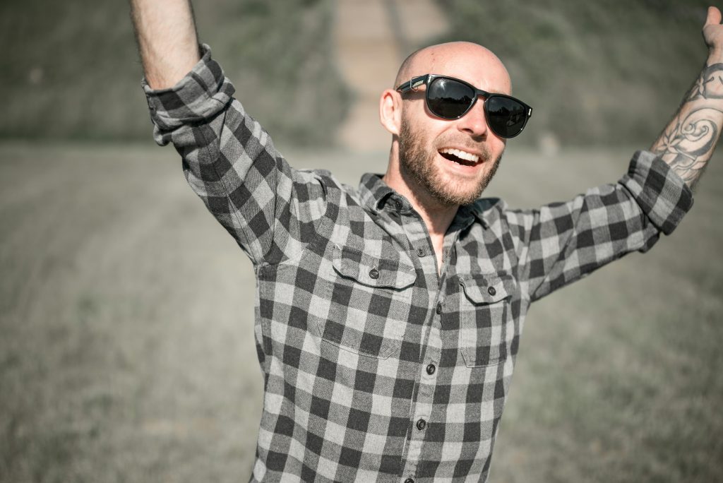 Bald man with sunglasses raises arms joyfully outdoors, exuding happiness.